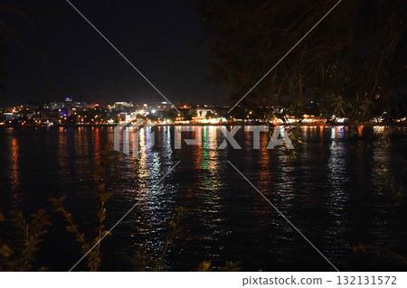 A Serene Night by the Water, Capturing the Sparkling Reflections of City Lights on Calm Waters, Surrounded by Lush Greenery and a Peaceful Atmosphere A Serene Night by the Water, Capturing the Sparkling Reflections of City Lights on Calm Waters, Surrounded by Lush Greenery and a Peaceful Atmosphere 132131572
