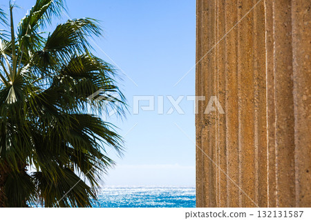 A Serene Coastal View with Palm Trees Framing a Stone Column Against a Bright Blue Sky and Sparkling Ocean Waves in the Background A Serene Coastal View with Palm Trees Framing a Stone Column Against a Bright Blue Sky and Sparkling Ocean Waves in the Background 132131587