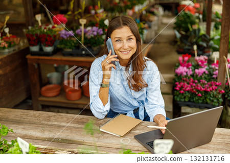 A Young Woman Engaged in Productive Work in a Colorful Garden Shop, Using Her Laptop and Phone 132131716