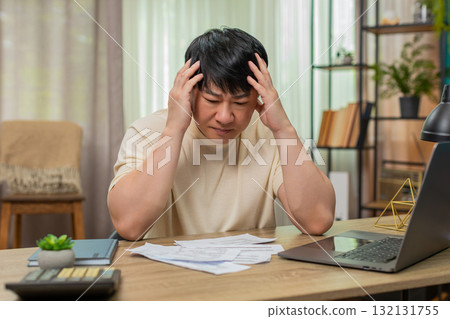 Asian man holding head in stress, surrounded by unpaid bills on table, calculator and laptop nearby 132131755
