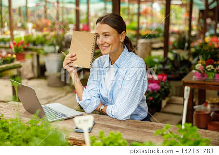 A woman is actively engaged in her work at a flower shop, using a laptop and notepad to assist her A woman is actively engaged in her work at a flower shop, using a laptop and notepad to assist her 132131781