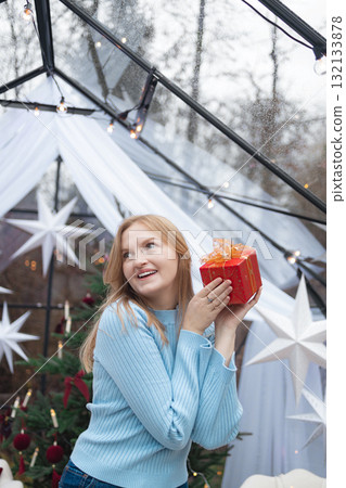 Smiling blonde Caucasian woman holding red Christmas gift near face in festive setting Smiling blonde Caucasian woman holding red Christmas gift near face in festive setting 132133878