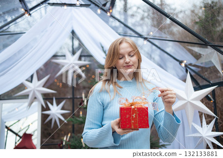 Blonde Caucasian woman opening red Christmas gift box in festive setting 132133881