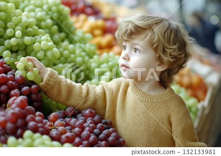 Little child in a warm sweater picking green grapes at a vibrant fruit market surrounded by colorful bunches of fresh grapes 132133981