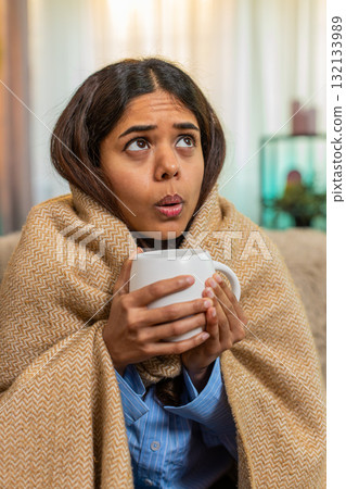 Indian woman drinking hot tea and coughing at home under blanket, showing cold and tired expression Indian woman drinking hot tea and coughing at home under blanket, showing cold and tired expression 132133989