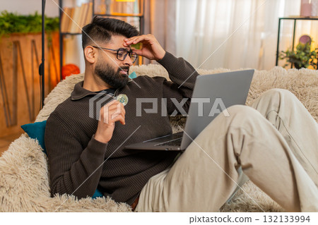 Indian man looks disappointed while closing laptop, reacting to Bitcoin price drop on sofa at home 132133994