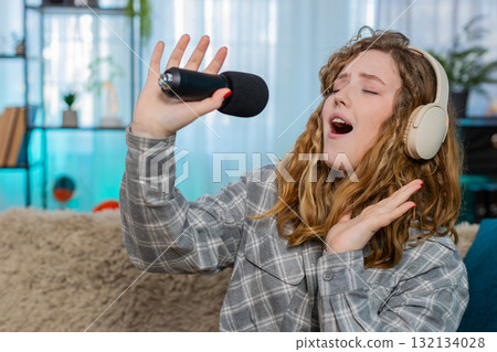 Woman on sofa singing with microphone wearing headphones moving joyfully to music feeling like star Woman on sofa singing with microphone wearing headphones moving joyfully to music feeling like star 132134028