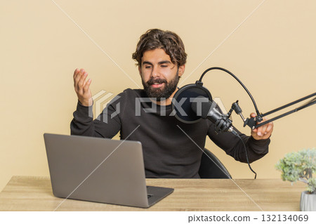 Happy blogger man recording podcast speaking into microphone and laptop webcam on beige background Happy blogger man recording podcast speaking into microphone and laptop webcam on beige background 132134069