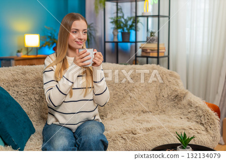 Caucasian woman enjoying hot tea on sofa holding cup both hands relaxing indoors calm comfortable 132134079