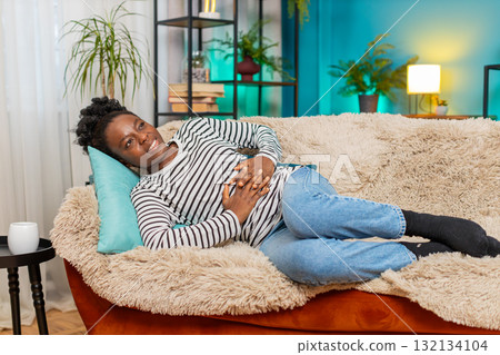 African American woman feeling stomach pain, holding abdomen, lying exhausted on couch indoors African American woman feeling stomach pain, holding abdomen, lying exhausted on couch indoors 132134104