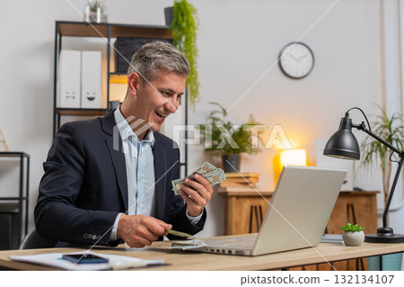 Caucasian young businessman in suit working on laptop PC counting money cash at home office desk 132134107