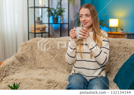 Caucasian woman enjoying hot tea on sofa holding cup both hands relaxing indoors calm comfortable 132134144