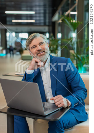 Mature businessman in suit thinking and dreaming while using laptop sitting in modern office lobby Mature businessman in suit thinking and dreaming while using laptop sitting in modern office lobby 132134170