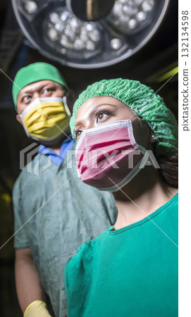 Asian male surgeon and latina nurse preparing for complex operation, wearing protective masks, gloves and scrub attire. Bright operating room lights. Asian male surgeon and latina nurse preparing for complex operation, wearing protective masks, gloves and scrub attire. Bright operating room lights. 132134598