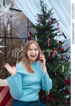 Smiling blonde Caucasian woman holding red mug near Christmas tree in cozy festive interior Smiling blonde Caucasian woman holding red mug near Christmas tree in cozy festive interior 132135024