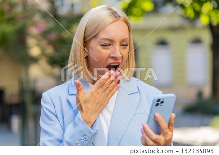Caucasian mature businesswoman looking shocked while reading good news on smartphone on city street 132135093