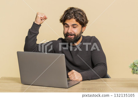 Happy man sitting at table using laptop winning lottery and celebrating success on beige background 132135105
