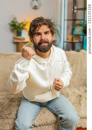 Happy young man watching TV celebrating success, winning, lottery luck, victory sitting on sofa 132135108