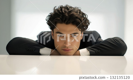 Handsome young man in black suit leaning on white table with intense gaze in modern light interior 132135120