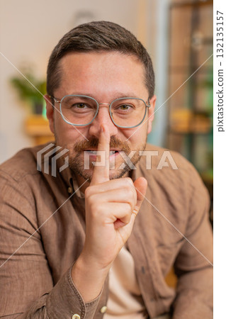 Caucasian young businessman presses index finger to lips makes silence gesture at home office table 132135157