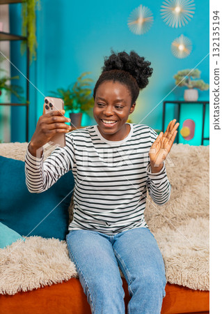 African American woman smiling waving smartphone hand gesture greeting happy indoors sofa African American woman smiling waving smartphone hand gesture greeting happy indoors sofa 132135194