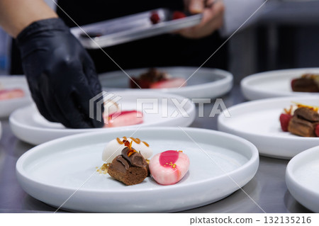 plated desserts in professional restaurant kitchen. chef arranges chocolate cake, mousse, and artistic garnishes on plates. fine dining presentation, culinary art and gourmet food preparation. 132135216