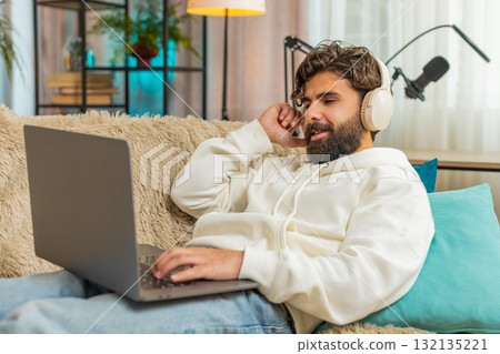 Happy relaxed Indian young man in wireless headphones listening to music using laptop on sofa couch Happy relaxed Indian young man in wireless headphones listening to music using laptop on sofa couch 132135221