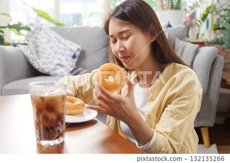 Happy young Asian woman enjoying a sweet glazed donut and iced cola at home, happy smiling in relaxation, showing simple joy and comfort food pleasure in a cozy lifestyle setting Happy young Asian woman enjoying a sweet glazed donut and iced cola at home, happy smiling in relaxation, showing simple joy and comfort food pleasure in a cozy lifestyle setting 132135266
