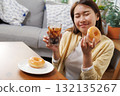 Happy young Asian woman enjoying a sweet glazed donut and iced cola at home, happy smiling in relaxation, showing simple joy and comfort food pleasure in a cozy lifestyle setting 132135267