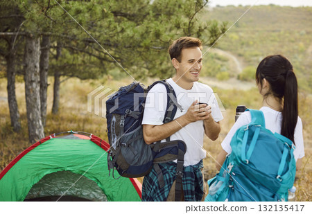Smiling hikers share drink by tent, forest campsite during sunset weekend 132135417