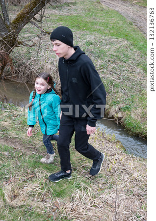 Exploring nature together on a cool day by the flowing stream, a young boy enjoys a delightful walk with his little sister surrounded by lush greenery and gentle sunshine Exploring nature together on a cool day by the flowing stream, a young boy enjoys a delightful walk with his little sister surrounded by lush greenery and gentle sunshine 132136763