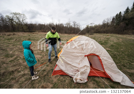 Family bonding experience in a tranquil campsite as a child watches with joy while an adult sets up a tent on a cloudy day in the heart of nature Family bonding experience in a tranquil campsite as a child watches with joy while an adult sets up a tent on a cloudy day in the heart of nature 132137064