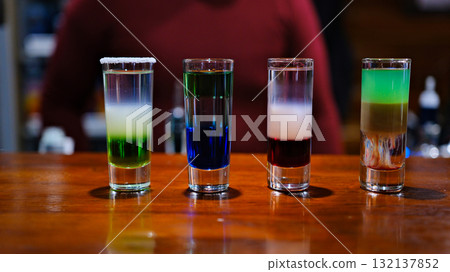 shot glasses filled with vibrant and colorful drinks sits on a sleek bar counter. The various hues reflect light and create an energetic atmosphere, perfect for a night out. 132137852