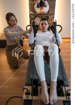 Woman Pilates Instructor Gives Guidance to a men Student While They Utilize the Reformers Equipment Woman Pilates Instructor Gives Guidance to a men Student While They Utilize the Reformers Equipment 132139745