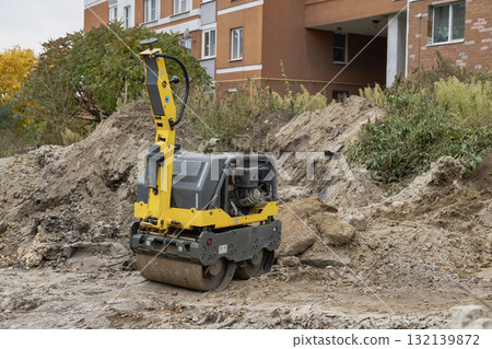 Construction site with a small vibrating roller compacting dirt near residential buildings in autumn Construction site with a small vibrating roller compacting dirt near residential buildings in autumn 132139872