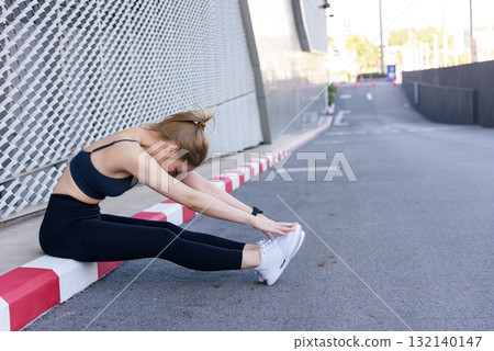 Workout woman performs outdoor sport exercise, stretching fit body for flexibility, enjoying active workout on urban street curb beside modern building for healthy lifestyle. 132140147