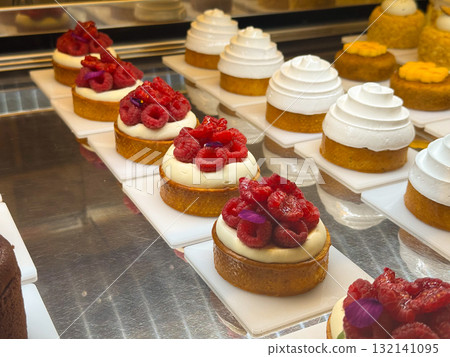 Round pastries with white meringue topping and cakes decorated with red berries arranged on white plates. Bakery display combining cream, color and craft. 132141095