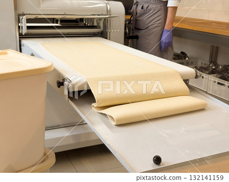 Layer of dough passing through rolling machine in bakery. Culinary preparation showing craft, precision, and texture of handmade baking. Layer of dough passing through rolling machine in bakery. Culinary preparation showing craft, precision, and texture of handmade baking. 132141159