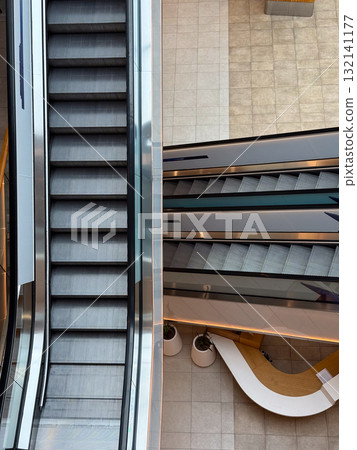 Escalators inside a contemporary shopping center viewed from above. Movement, symmetry, and urban architecture symbolizing daily routine and structure. Escalators inside a contemporary shopping center viewed from above. Movement, symmetry, and urban architecture symbolizing daily routine and structure. 132141177