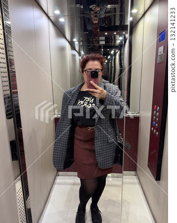 Woman taking mirror selfie in elevator wearing plaid blazer. Concept of urban self-expression, modern confidence, and visual symmetry in reflective architectural environment. 132141233