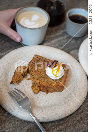 A slice of carrot cake with cream cheese frosting, pecan, and orange zest served with a cup of latte in a cozy cafe 132142046