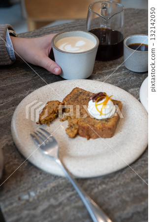 A slice of carrot cake with cream cheese frosting, pecan, and orange zest served with a cup of latte in a cozy cafe 132142050