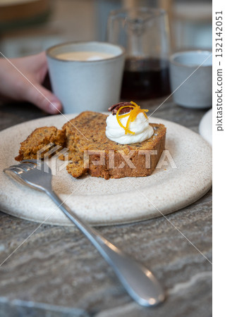 A slice of carrot cake with cream cheese frosting, pecan, and orange zest served with a cup of latte in a cozy cafe 132142051