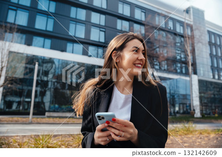 Young Woman Smiles While Using Smartphone Outside Modern Building in Urban Setting During Daylight Hours Young Woman Smiles While Using Smartphone Outside Modern Building in Urban Setting During Daylight Hours 132142609