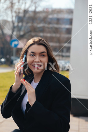 Smiling Young Woman Talking on the Phone Outdoors in a City Environment During Daytime With Clear Skies 132142621