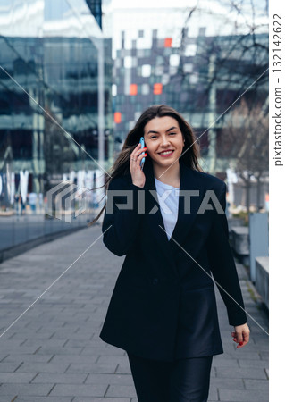 Professional Woman Walking Down City Sidewalk While Talking on Her Smartphone, Dressed in Stylish Black Blazer and Casual White Shirt on a Sunny Day Professional Woman Walking Down City Sidewalk While Talking on Her Smartphone, Dressed in Stylish Black Blazer and Casual White Shirt on a Sunny Day 132142622