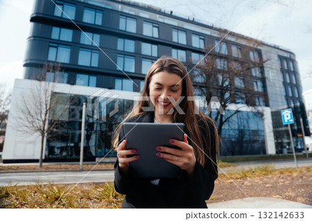Young woman enjoying digital tablet outdoors in urban setting with modern architecture and greenery surrounding her, technology lifestyle concept 132142633