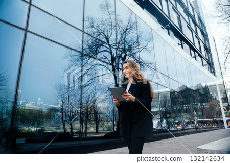 Professional woman walking with tablet near modern glass building reflecting trees and city life showcasing urban lifestyle concept Professional woman walking with tablet near modern glass building reflecting trees and city life showcasing urban lifestyle concept 132142634