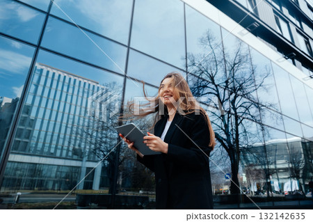 Confident businesswoman smiling while holding tablet outdoors near modern glass building reflecting trees and sky, professional lifestyle concept Confident businesswoman smiling while holding tablet outdoors near modern glass building reflecting trees and sky, professional lifestyle concept 132142635