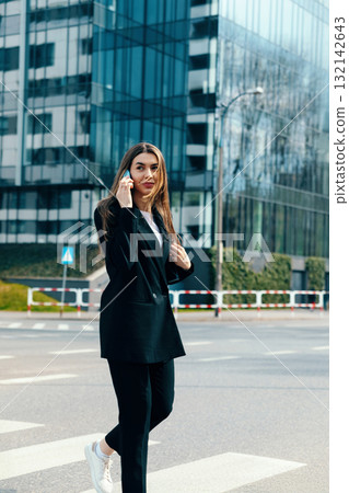 Confident young woman in stylish black suit walking on city street with modern buildings in background showcasing urban fashion and lifestyle concept 132142643